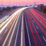 Langzeitbelichtung einer Autobahn bei Dämmerung mit leuchtenden Lichtspuren von Fahrzeugen in Weiß und Rot, die eine dynamische Bewegung auf den mehrspurigen Fahrbahnen darstellen.